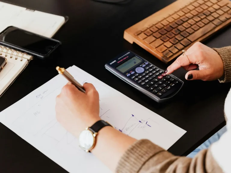 Close-up of hands working with a calculator and notebook on a desk, analyzing documents.